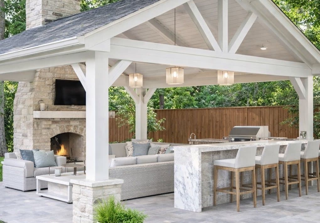 Covered outdoor living pavilion with painted timber trusses and a tongue-and-groove ceiling, a full-height stone fireplace with a timber mantel, and a marble-clad outdoor kitchen with a built-in grill and bar seating